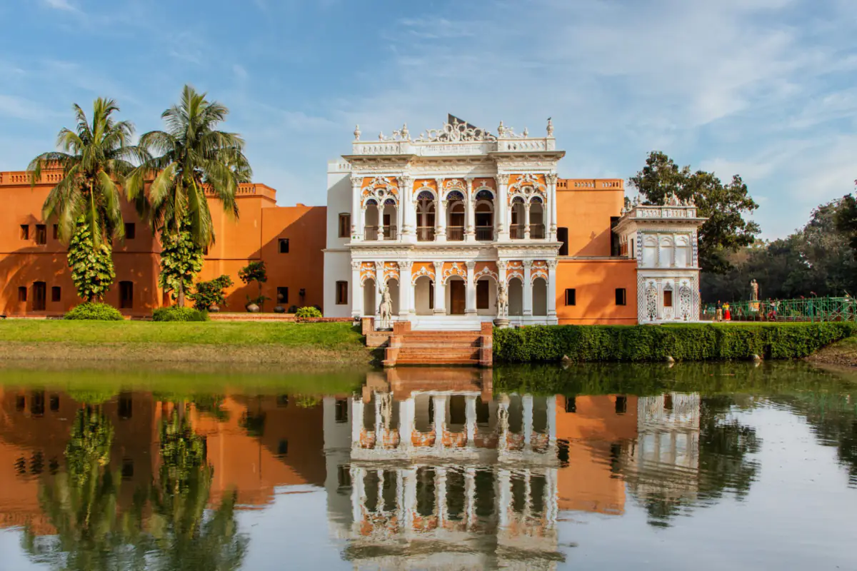 An ornate white and orange mansion with palm trees beside it, reflected in a still pond