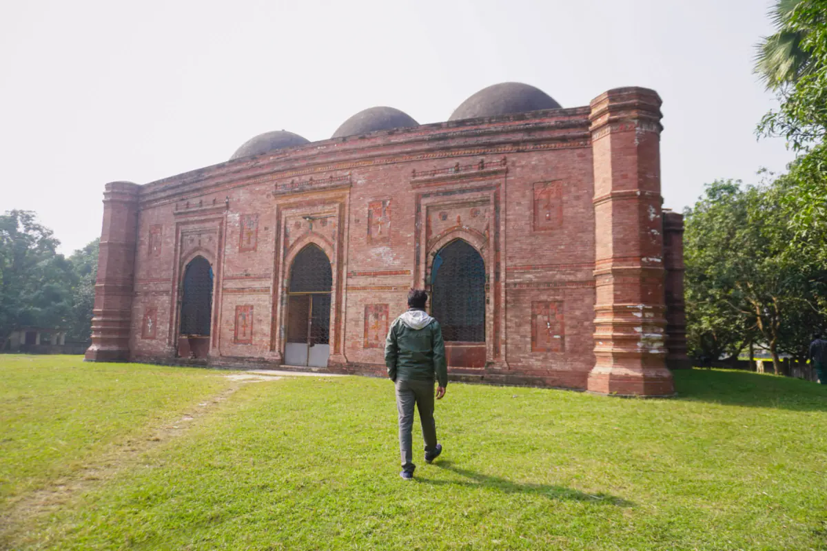A visitor walking across a lawn towards an old red brick mosque with three domes in Bagerhat